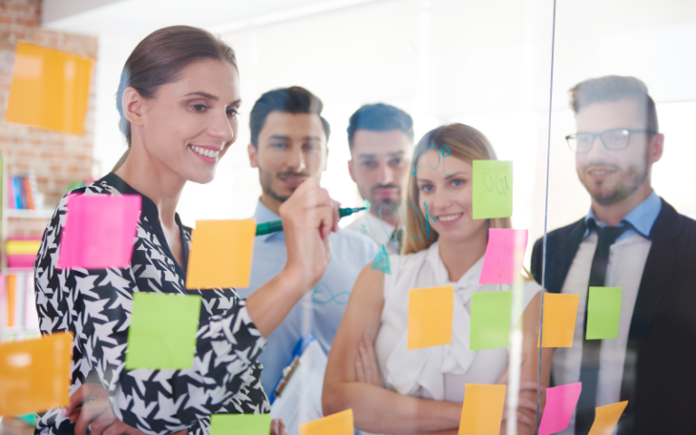 Na imagem, vemos um grupo de profissionais observando post-its coloridos colados em uma parede de vidro. O grupo está sorridente ao observar seu desenvolvimento e resultados. A ideia é representar os motivos de aplicar o pdi na sua empresa.