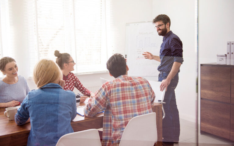 A imagem mostra um grupo de pessoas animadas conversando em uma sala. Ao fundo, observa-se um quadro branco com gráficos e dados. Eles conversam sobre estruturar um plano de desenvolvimento de carreira na empresa em que trabalham.