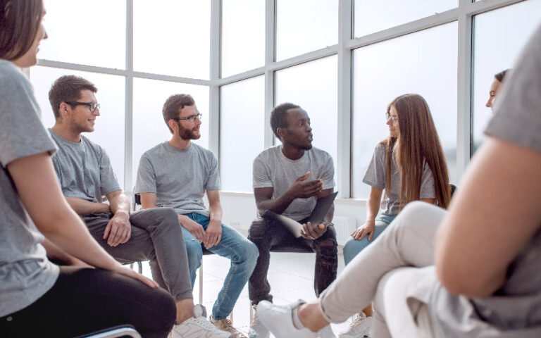 Dinâmica de grupo sendo realizada por sete pessoas jovens e diversas. Elas disputam uma vaga em um importante processo seletivo.