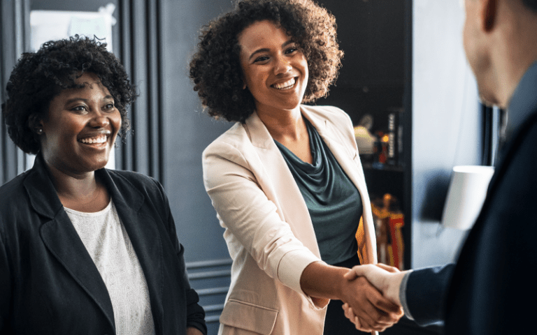 Imagem de capa para blog post sobre tipos de recrutamento. Duas mulheres sorridentes em um ambiente corporativo, enquanto uma delas aperta a mão de um homem de terno.