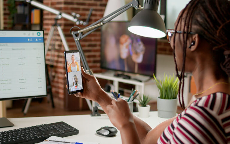 Imagem de mulher em chamada de vídeo no celular. A sua frente, está um computador com documentos abertos. A jovem sorri, representando uma boa Candidate Experience no processo seletivo.
