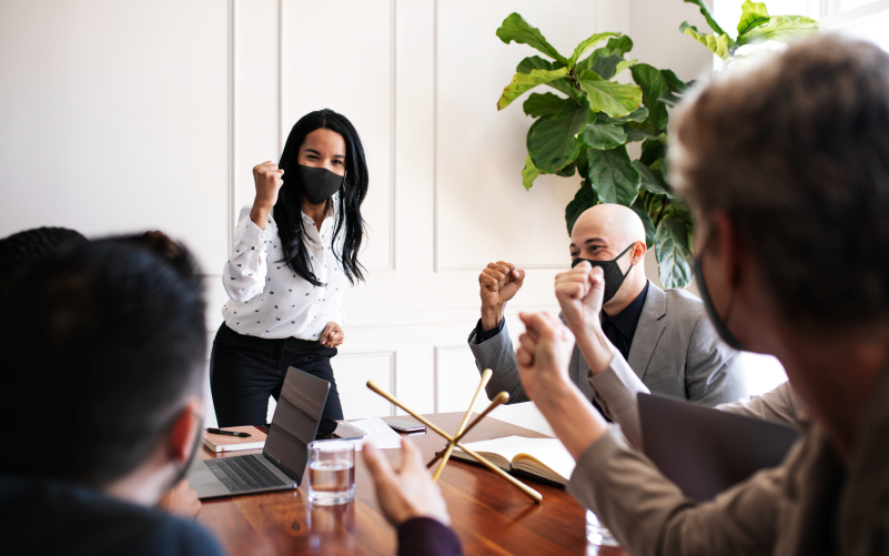 Imagem de equipe sentada ao redor de uma mesa, representando uma cultura participativa. Em destaque, está uma mulher em pé, de forma a representar a figura da liderança.