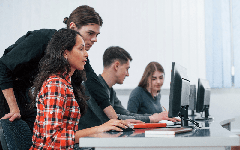 Imagem de capa para artigo sobre como contratar profissionais de TI. Equipe de tecnologia trabalha em conjunto em uma bancada de computadores. Em destaque, um desenvolvedor auxilia uma colega na resolução de um problema de programação na tela do monitor.