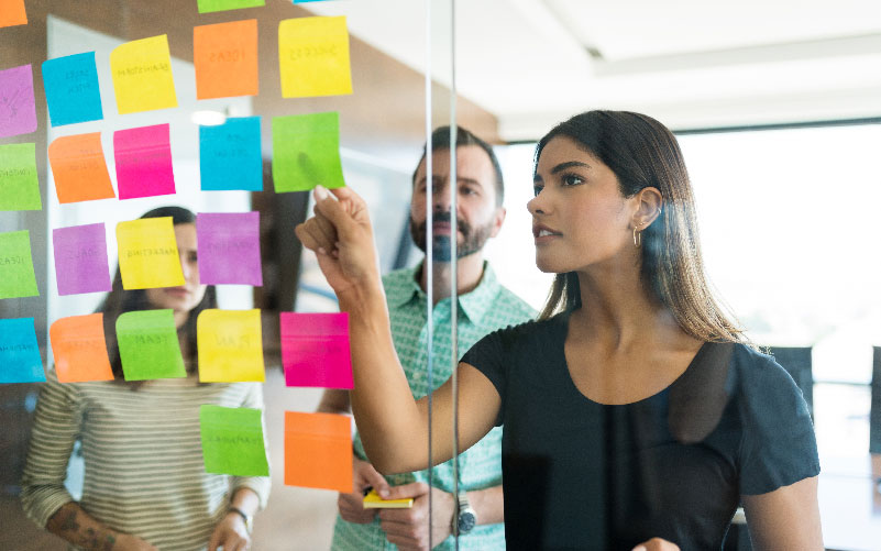 A imagem traz um vidro em primeiro plano. Nele, estão colados post-its coloridos e uma mulher apresenta seus conteúdos para um grupo. A imagem busca representar o uso de design thinking no RH.