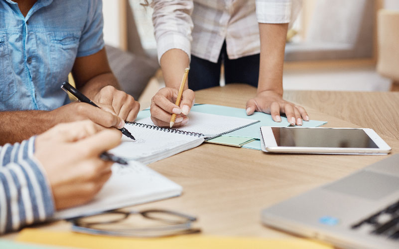 A imagem foca nas mãos de três pessoas, enquanto elas trabalham. Na mesa em que se apoiam, aparecem papéis, um tablet, parte de um notebook e um óculos. O objetivo é representar o planejamento de ações de endomarketing nas empresas.