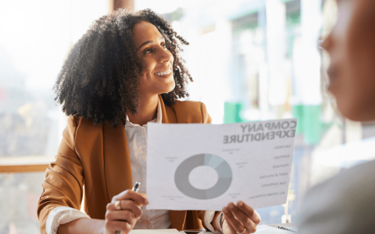 Imagem de capa para artigo sobre eNPS. Na imagem, mostra uma mulher afrodescendente sorrindo em reunião de trabalho, segurando um gráfico e um relatório impresso com dados, enquanto interage com colegas ao fundo.