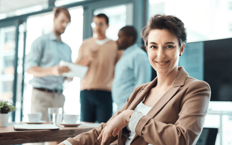 Imagem de capa para artigo sobre Liderança Positiva. Na imagem, mostra uma mulher sorridente em primeiro plano, usando jaleco e posando ao lado de uma mesa em ambiente corporativo, enquanto colegas conversam ao fundo em escritório moderno.