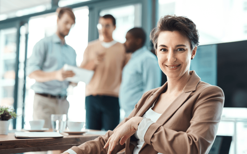 Imagem de capa para artigo sobre Liderança Positiva. Na imagem, mostra uma mulher sorridente em primeiro plano, usando jaleco e posando ao lado de uma mesa em ambiente corporativo, enquanto colegas conversam ao fundo em escritório moderno.