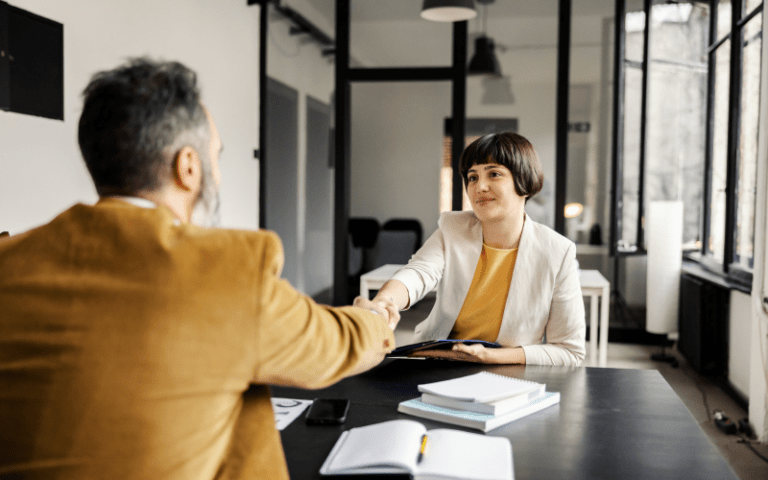 A imagem mostra uma gestora oferecendo um aperto de mão para uma entrevista para atração de um talento para sua empresa.