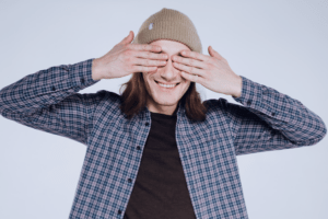 Homem jovem com as mãos no rosto, tampando os olhos. A imagem busca retratar, de forma lúdica, a dinâmica “trilha com olhos vendados”, que pode ser aplicada no team building.