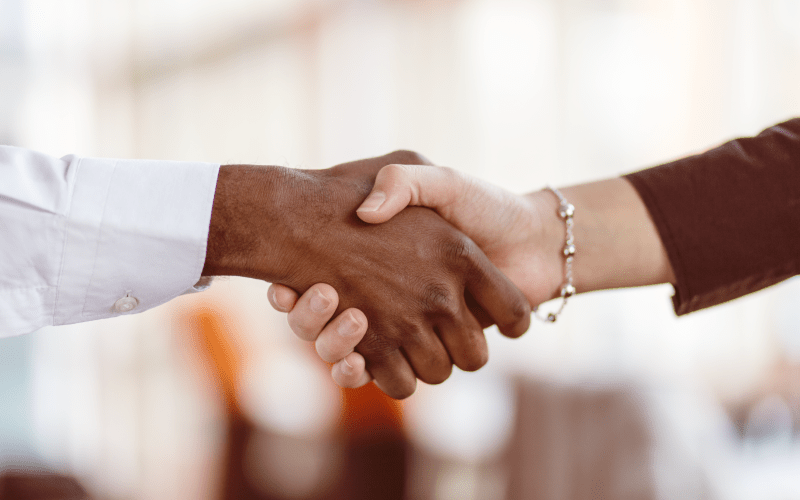 Duas pessoas apertam as mãos em um ambiente corporativo, simbolizando a formalização de um acordo profissional. A imagem ilustra o processo de contratação e os diferentes tipos de contrato de trabalho.