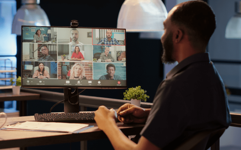 Imagem de capa para artigo sobre E-learning. Homem visto de costas participa de uma videochamada em um monitor grande com vários outros participantes, ilustrando uma aula ou treinamento online.