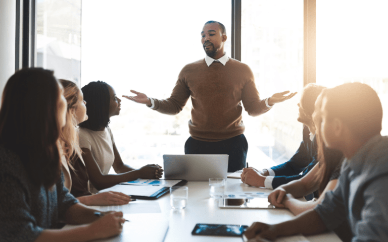 Um homem está de pé, conduzindo uma conversa com cinco colegas sentados ao redor de uma mesa de reunião, cada um com um notebook ou caderno, em um escritório corporativo. A imagem ilustra um contexto de diálogo e colaboração entre equipes, relacionado à gestão de conflitos no ambiente de trabalho.