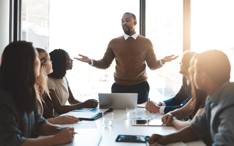 Um homem está de pé, conduzindo uma conversa com cinco colegas sentados ao redor de uma mesa de reunião, cada um com um notebook ou caderno, em um escritório corporativo. A imagem ilustra um contexto de diálogo e colaboração entre equipes, relacionado à gestão de conflitos no ambiente de trabalho.