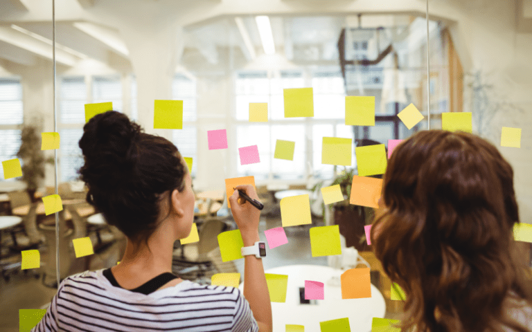Duas profissionais organizam ideias em um quadro de vidro com post-its coloridos em um escritório. A imagem representa uma dinâmica colaborativa de planejamento e priorização de atividades, comum na metodologia Sprint.