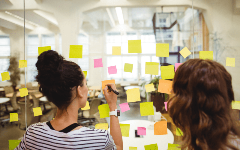 Duas profissionais organizam ideias em um quadro de vidro com post-its coloridos em um escritório. A imagem representa uma dinâmica colaborativa de planejamento e priorização de atividades, comum na metodologia Sprint.