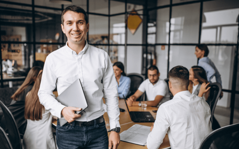 Homem sorridente vestindo camisa social branca, segurando um notebook fechado e olhando para a câmera, representando o profissional 5.0 que alia tecnologia e humanização. Ao fundo, sua equipe colabora ao redor de uma mesa de reunião em um escritório moderno.