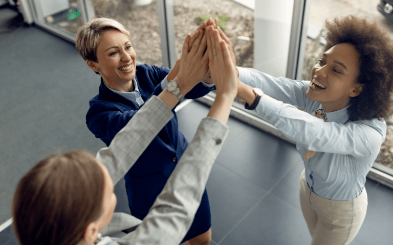 Imagem de capa para artigo sobre como construir relações de confiança no trabalho. Três profissionais mulheres sorriem e celebram juntas, unindo as mãos no alto em um gesto de parceria e trabalho em equipe, vistas por um ângulo superior dentro do escritório.