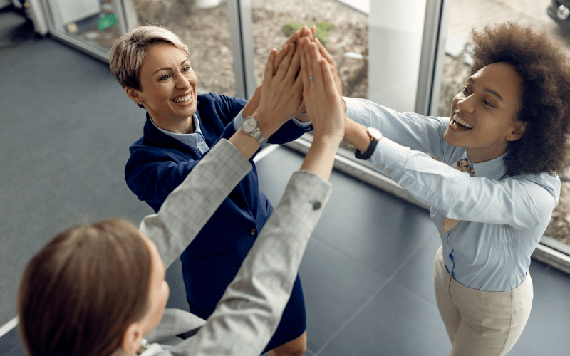 Imagem de capa para artigo sobre como construir relações de confiança no trabalho. Três profissionais mulheres sorriem e celebram juntas, unindo as mãos no alto em um gesto de parceria e trabalho em equipe, vistas por um ângulo superior dentro do escritório.