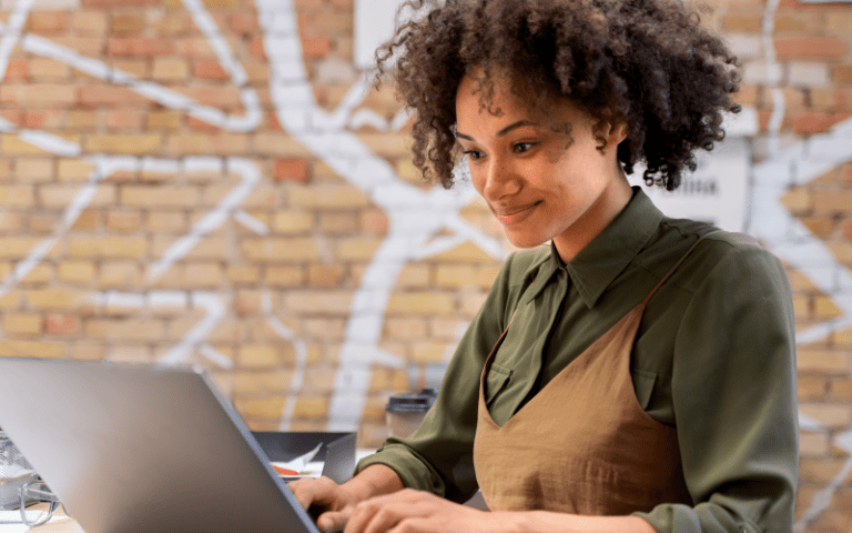 Uma jovem profissional negra sorrindo enquanto usa um notebook em um escritório com parede de tijolos expostos. A imagem representa a gestão de benefícios sendo feita de maneira estratégica.