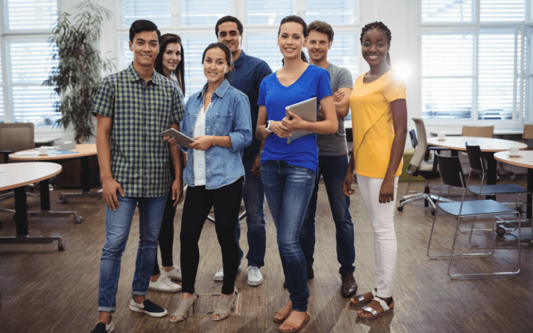 Um grupo diversificado de jovens profissionais está em pé, em um escritório moderno de plano aberto, sorrindo e olhando para a câmera. Duas mulheres seguram tablets, enquanto os outros estão relaxados. A imagem representa uma equipe unida e o sucesso da gestão de talentos.