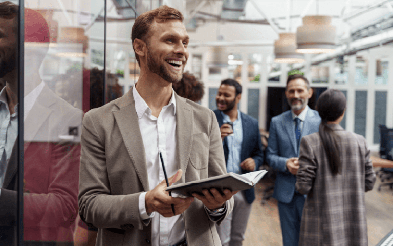 Em um escritório corporativo, um profissional sorri enquanto segura um tablet e conversa com colegas durante uma interação de trabalho. Ao fundo, outros colaboradores conversam em um ambiente moderno e colaborativo, ilustrando práticas de boa liderança, comunicação e engajamento entre equipes.