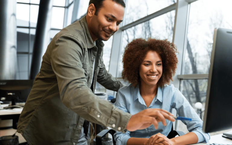 Dois profissionais autônomos colaboram de forma dinâmica diante da tela de um computador em um moderno espaço de coworking. Uma mulher negra com cabelos crespos volumosos sorri e digita sentada, enquanto um homem de jaqueta verde se inclina ao seu lado apontando para o monitor. A interação retrata a flexibilidade, a colaboração por projetos e a autonomia características da gig economy no mercado de trabalho atual.