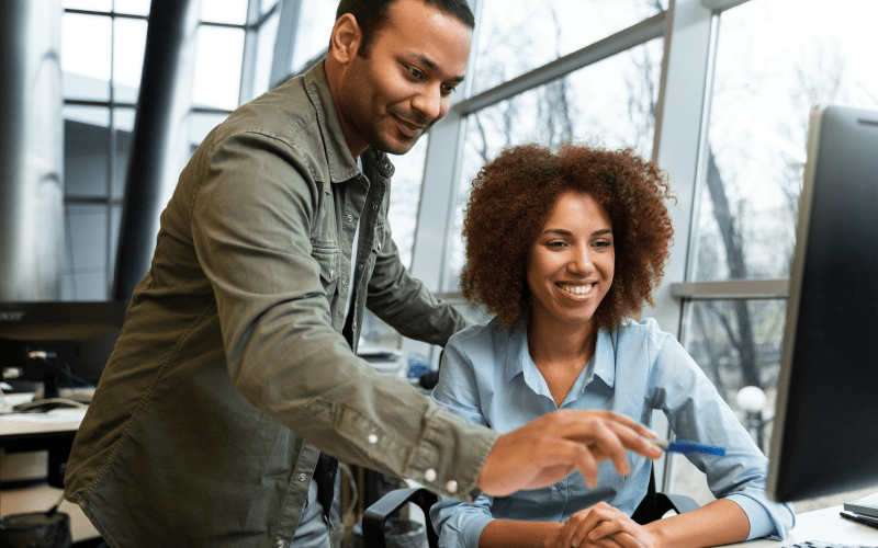 Dois profissionais autônomos colaboram de forma dinâmica diante da tela de um computador em um moderno espaço de coworking. Uma mulher negra com cabelos crespos volumosos sorri e digita sentada, enquanto um homem de jaqueta verde se inclina ao seu lado apontando para o monitor. A interação retrata a flexibilidade, a colaboração por projetos e a autonomia características da gig economy no mercado de trabalho atual.