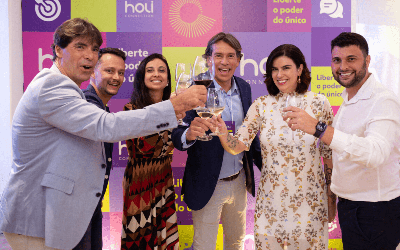 Grupo de seis profissionais brindando com taças de vinho em evento, à frente de painel com a marca Holi Connection e a frase 'Liberte o poder do único'.