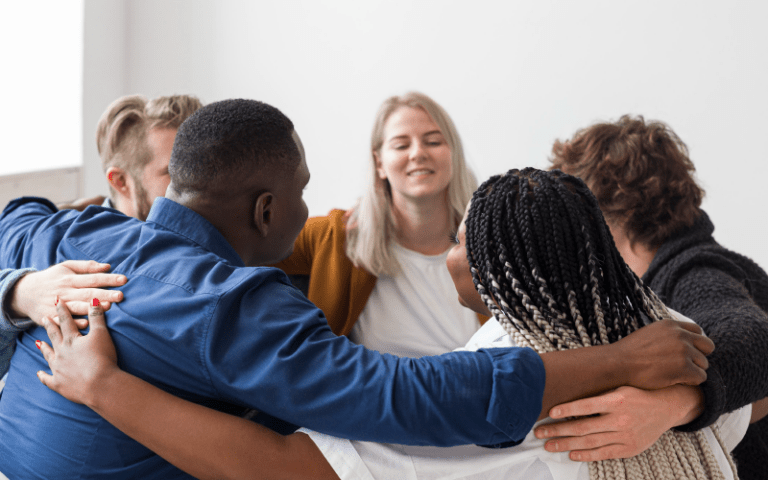 Grupo diverso de pessoas em círculo, abraçadas pelos ombros, sorrindo em um momento de acolhimento e união em ambiente de trabalho, representando um recrutamento inclusivo.