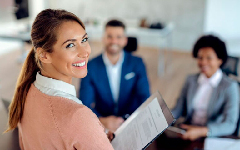 Imagem de capa de post blog sobre recrutamento temporário nas empresas. Na foto, temos três pessoas em um ambiente de trabalho. Observamos uma mulher em foco sorrindo, enquanto as duas outras pessoas estão desfocadas, em segundo plano.