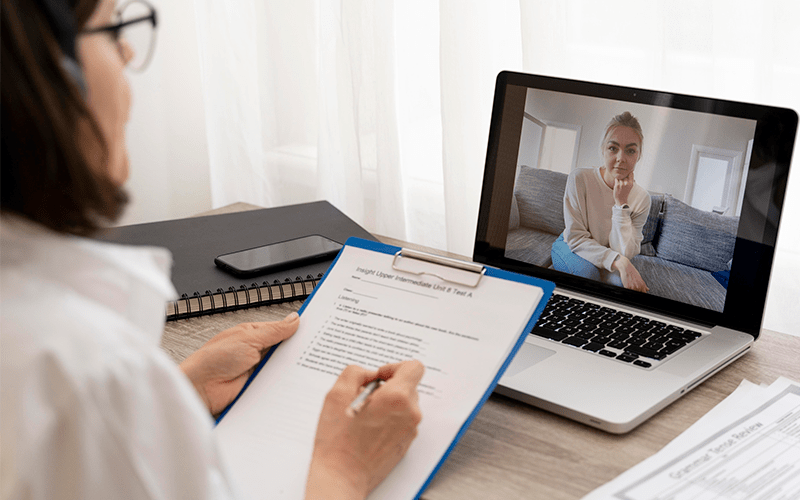 Recrutadora realiza entrevista online e avaliação de proficiência com uma candidata estrangeira por videochamada no notebook. Em primeiro plano, a profissional acompanha o processo segurando uma prancheta com um teste de inglês, enquanto a candidata aparece na tela do computador direto de sua casa, representando a flexibilidade, a tecnologia e a conexão global na contratação internacional de talentos.