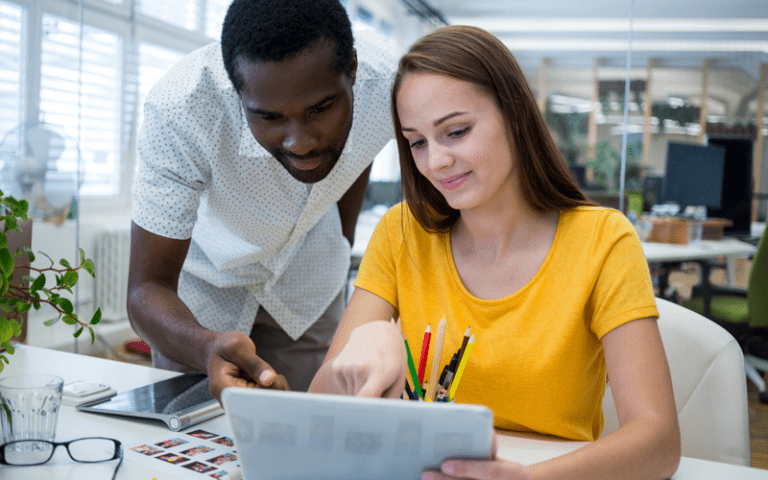 Dois profissionais colaboram de forma próxima e integrada sobre um tablet em um escritório moderno. Um homem negro de camisa estampada se inclina, apontando para a tela, enquanto uma mulher branca de camiseta amarela observa com atenção. A interação demonstra a troca de conhecimento multidisciplinar e a colaboração horizontal, características fundamentais de uma equipe composta por profissionais T-shaped.