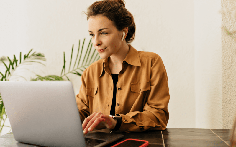 Recrutadora trabalha de forma focada diante de seu notebook, realizando a busca ativa e a triagem de candidatos qualificados. Vestindo uma camisa mostarda e fones de ouvido sem fio, ela exibe uma expressão de atenção e satisfação, representando a eficiência e a estratégia do processo de recrutamento pelo LinkedIn em um ambiente moderno.