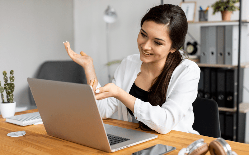 Recrutadora sorri com empatia e gesticula de forma acolhedora durante uma videochamada no notebook, transmitindo um feedback de entrevista humanizado para um candidato. Ela veste blusa social branca e está em um escritório organizado, representando a importância de uma comunicação clara, transparente e positiva nos processos seletivos.