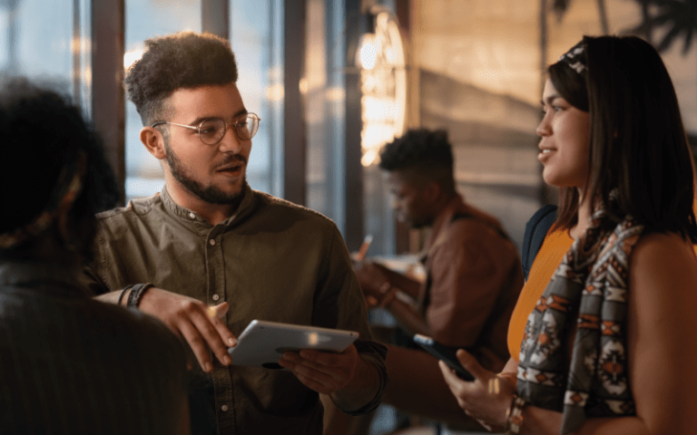 Três jovens profissionais conversam de forma atenta e colaborativa em um ambiente descontraído de co-working ou cafeteria. Um jovem negro de óculos e camisa verde, segurando um tablet, fala enquanto gesticula suavemente, demonstrando comunicação assertiva, enquanto uma mulher de blusa laranja o escuta com atenção, ilustrando empatia e habilidades socioemocionais na prática.
