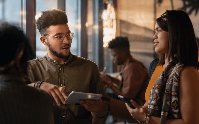 Três jovens profissionais conversam de forma atenta e colaborativa em um ambiente descontraído de co-working ou cafeteria. Um jovem negro de óculos e camisa verde, segurando um tablet, fala enquanto gesticula suavemente, demonstrando comunicação assertiva, enquanto uma mulher de blusa laranja o escuta com atenção, ilustrando empatia e habilidades socioemocionais na prática.
