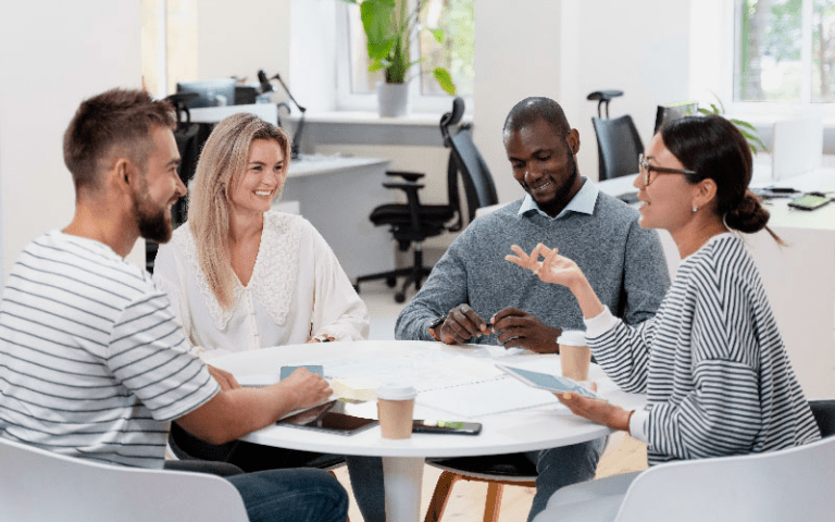 Equipe diversa com quatro profissionais sorridentes reunida ao redor de uma mesa redonda em um escritório compartilhado, representando a colaboração ativa, o pertencimento e a diversidade em uma empresa inclusiva.