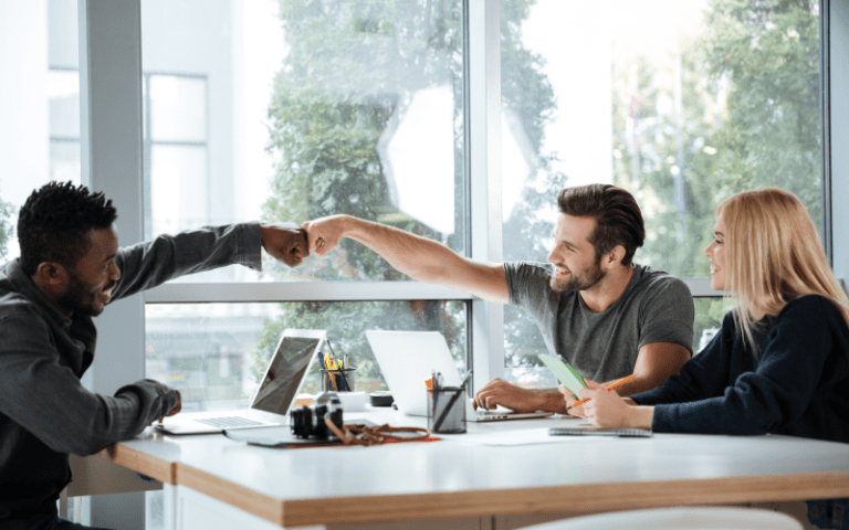 Um líder e seu liderado, dois homens de diferentes etnias, sorriem enquanto trocam um toque de punhos amigável sobre a mesa de trabalho, celebrando um progresso. Ao lado, uma colega loira acompanha o gesto com simpatia, ilustrando um ambiente corporativo saudável, colaborativo e pautado pela empatia e liderança humanizada.