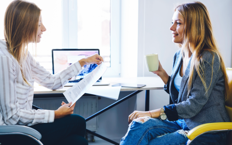 Duas profissionais conversam e sorriem em uma sessão de mentoria reversa em um escritório iluminado. À esquerda, uma jovem de blusa listrada entrega um documento impresso para a colega à direita, que veste blazer cinza e calça jeans rasgada enquanto segura uma caneca clara. A interação demonstra um ambiente colaborativo de aprendizado mútuo e troca de conhecimentos entre gerações.