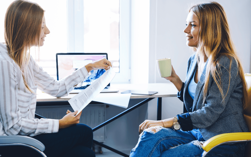 Duas profissionais conversam e sorriem em uma sessão de mentoria reversa em um escritório iluminado. À esquerda, uma jovem de blusa listrada entrega um documento impresso para a colega à direita, que veste blazer cinza e calça jeans rasgada enquanto segura uma caneca clara. A interação demonstra um ambiente colaborativo de aprendizado mútuo e troca de conhecimentos entre gerações.