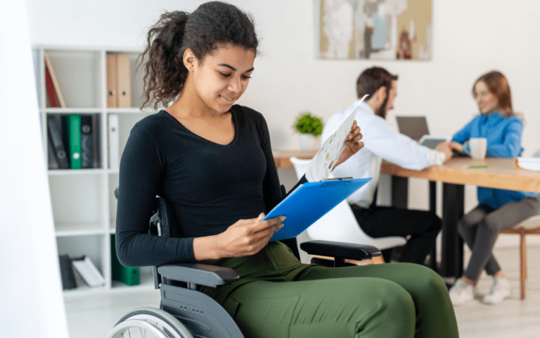 Profissional PcD, uma mulher negra cadeirante, analisa documentos de trabalho presos a uma prancheta azul com um leve sorriso. Ela está em primeiro plano em um escritório moderno, bem iluminado e acessível. Ao fundo e suavemente desfocados, dois colegas de trabalho conversam em uma mesa de reunião, representando um ambiente corporativo dinâmico e inclusivo.