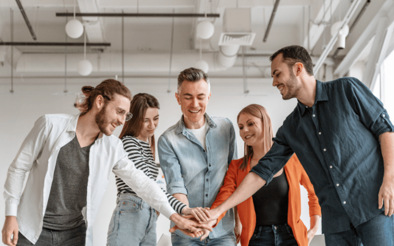 Equipe de cinco profissionais sorridentes une as mãos no centro em um gesto de união e colaboração. O grupo, composto por homens e mulheres vestidos de forma casual e descontraída, demonstra forte engajamento, sinergia e satisfação, representando uma excelente cultura de Employee Experience.