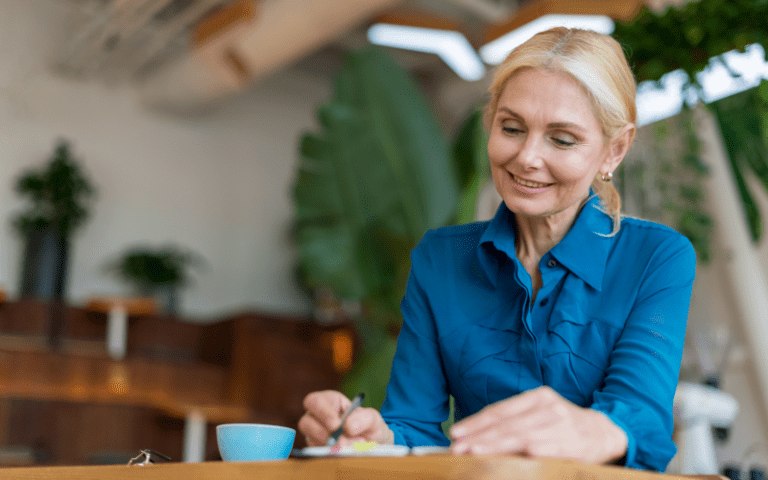 Uma profissional madura, mulher branca com cabelos loiros, trabalha com foco e satisfação em um escritório moderno. Vestindo camisa azul-turquesa, ela sorri enquanto faz anotações, representando a importância da diversidade geracional, da experiência e do combate ao etarismo no mercado de trabalho.