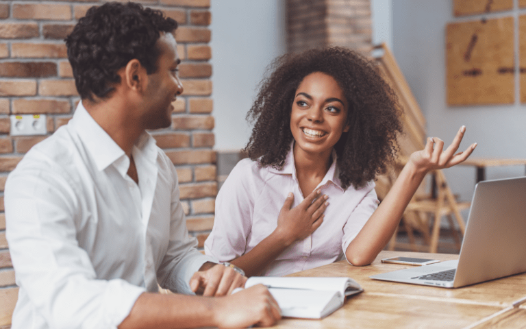 Dois profissionais em um momento de diálogo aberto e feedback contínuo no escritório. Uma mulher negra com cabelo crespo e camisa rosa sorri e gesticula de forma expressiva ao expor suas ideias. Ao seu lado, um homem de camisa branca a escuta com atenção e um semblante receptivo.
