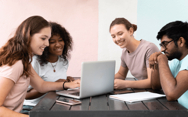 Quatro jovens de diferentes etnias colaboram e sorriem ao redor de um notebook sobre a mesa. A interação dinâmica e descontraída do grupo de nativos digitais representa a chegada e o comportamento colaborativo da Geração Alpha no ecossistema de trabalho do futuro.