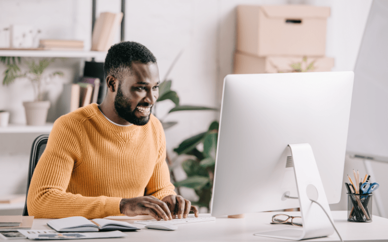 Profissional negro sorri com entusiasmo enquanto trabalha diante de um computador, representando a satisfação e o engajamento em uma jornada do colaborador positiva. Ele veste uma blusa de tricô mostarda e está em um escritório iluminado, acolhedor e moderno, com plantas ao fundo.