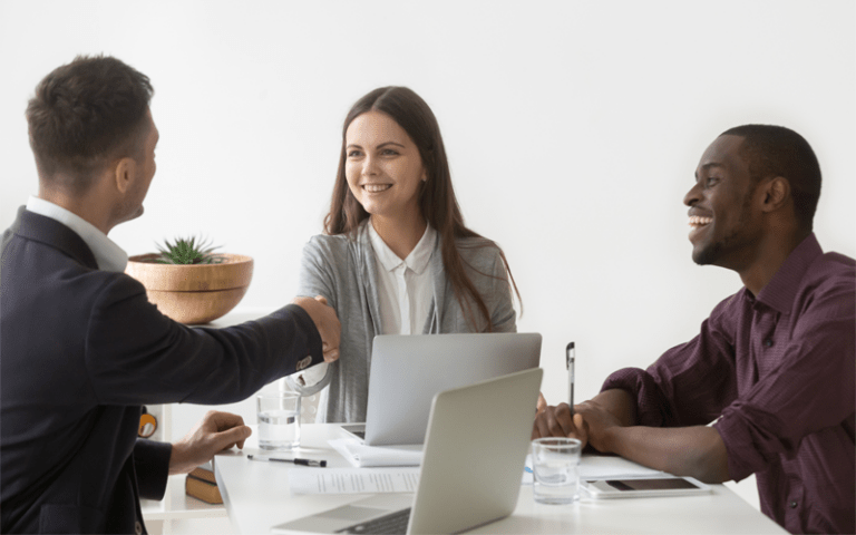 Uma recrutadora sorri ao fechar uma contratação de sucesso através de um aperto de mãos com o candidato ideal. Ao lado, um colega de trabalho negro ri de forma amigável, celebrando o momento. Eles estão sentados ao redor de uma mesa branca com notebooks e documentos em uma sala de entrevista iluminada.