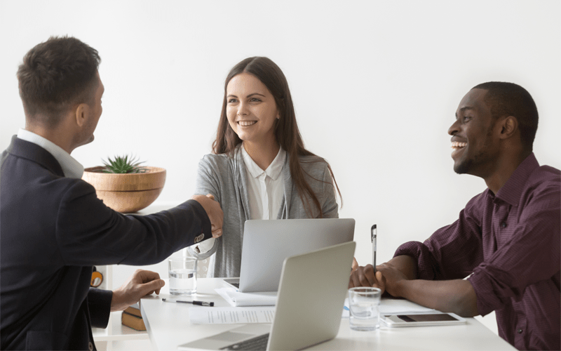 Uma recrutadora sorri ao fechar uma contratação de sucesso através de um aperto de mãos com o candidato ideal. Ao lado, um colega de trabalho negro ri de forma amigável, celebrando o momento. Eles estão sentados ao redor de uma mesa branca com notebooks e documentos em uma sala de entrevista iluminada.