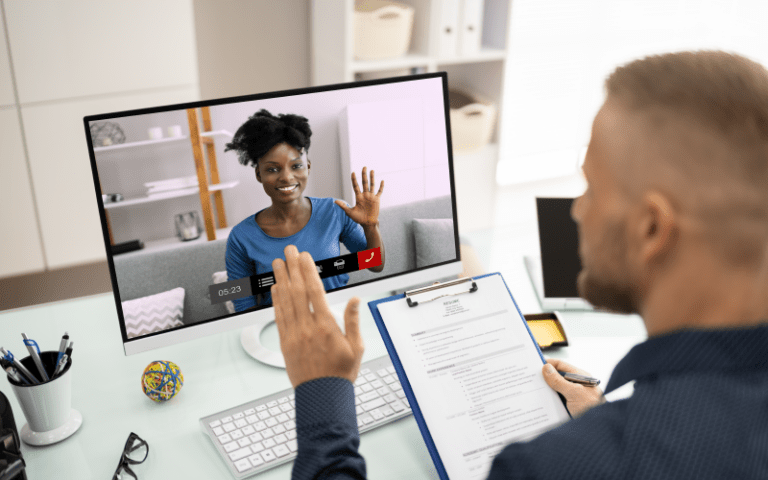 Recrutador conduz uma entrevista online estruturada seguindo um roteiro de entrevista e currículo impressos em sua prancheta azul. Pela perspectiva de trás do entrevistador, ele acena para a candidata, uma mulher negra que sorri e retribui o aceno na tela do monitor, demonstrando simpatia e conexão no processo seletivo.